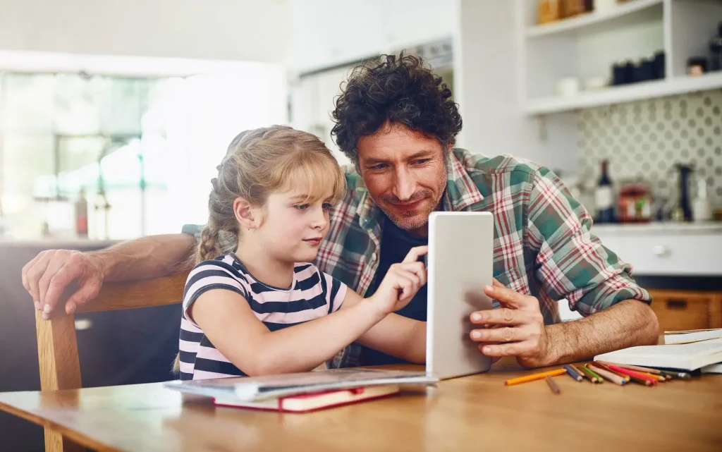 A man and a young girl look together at a tablet on a kitchen table with coloured pencils and books