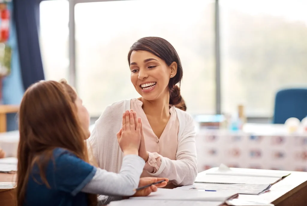 A support worker smiles and gives a high five to a young girl across a desk in a sunlit room