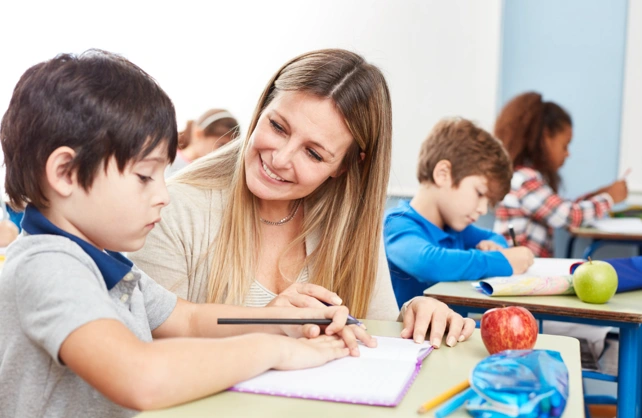 A teacher with long blonde hair helps a young boy writing in a notebook at a school desk