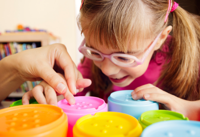 A young girl with pink glasses smiles while playing with colourful stacking cups on a table