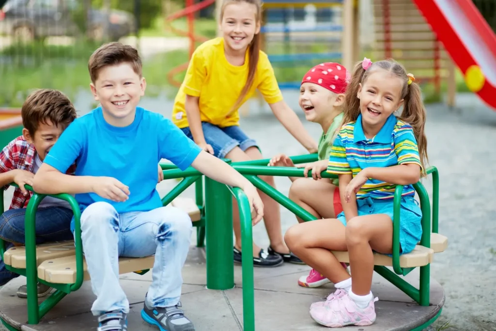 Five young children laughing and playing together on a green playground merry-go-round outdoors