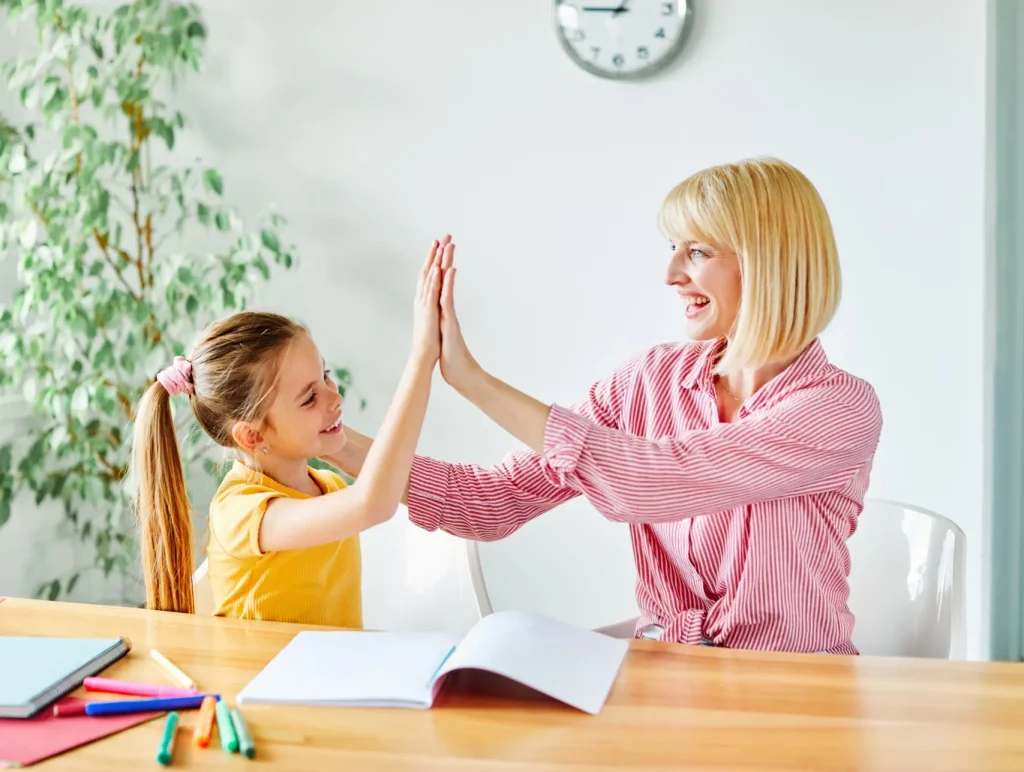 A woman gives a high five to a young girl at a wooden table with notebooks and coloured markers
