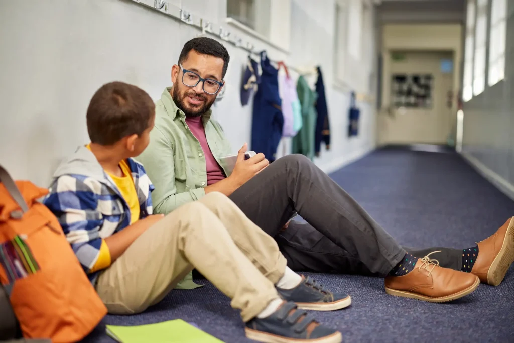 A support worker sits with a young boy in a school corridor, talking and writing in a notebook